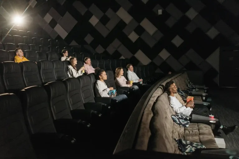 Pessoas assistindo a um filme em uma sala de cinema com assentos confortáveis, ambiente escuro e iluminação focada na tela.