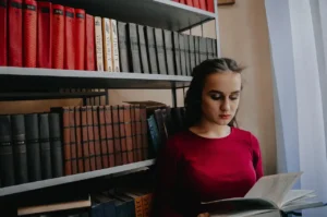 Jovem mulher lendo um livro na biblioteca com prateleiras cheias de livros ao fundo, destacando o momento de estudo e leitura.