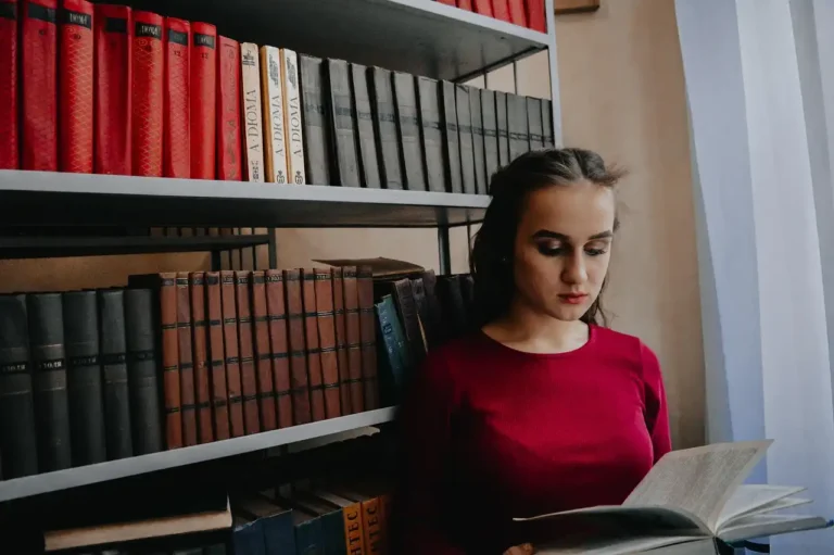 Jovem mulher lendo um livro na biblioteca com prateleiras cheias de livros ao fundo, destacando o momento de estudo e leitura.